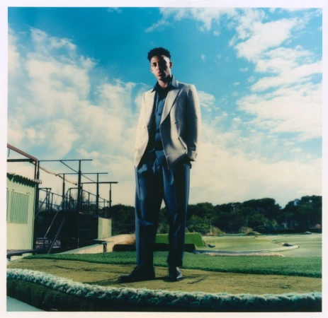 Alex Freeman posing outdoors on artificial turf in a cream blazer and navy blue trousers under a bright sky.