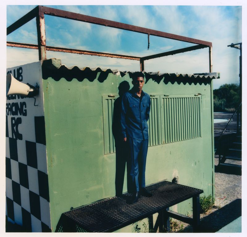 Alex Freeman in a blue matching suit standing on a metal platform beside a green racing track booth under bright sunlight.