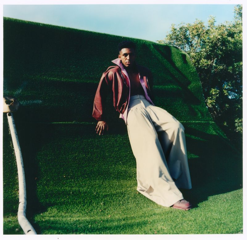 Alex Freeman wearing a burgundy leather jacket and wide-leg beige trousers resting on a green turf structure outdoors.