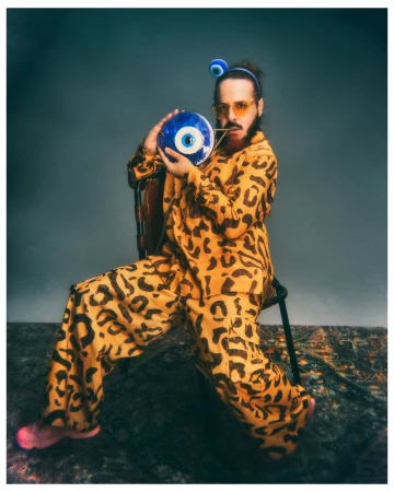 Man seated wearing a yellow leopard print suit holding a blue evil eye orb in front of a grey studio backdrop.