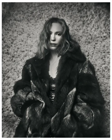 Black and white portrait of Hannah Einbinder wearing an oversized fur coat against a textured stone wall.