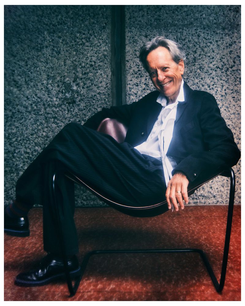 Richard E. Grant smiling while seated in a modern black sling chair against a textured concrete wall wearing a dark suit.