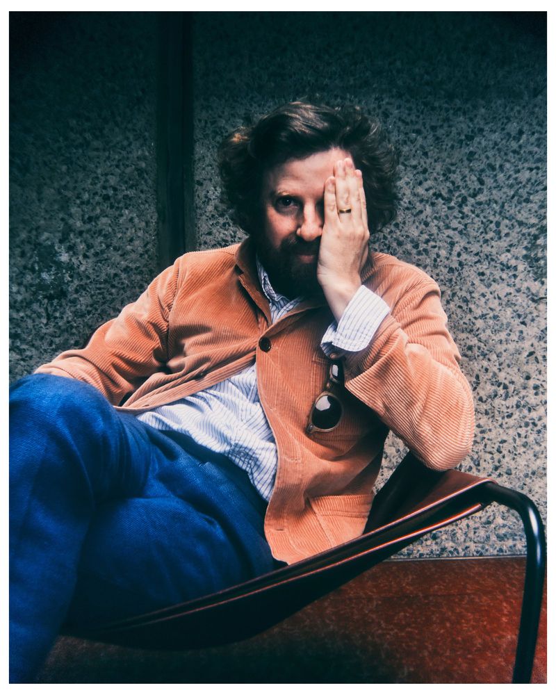 Man with beard wearing an orange corduroy jacket seated in a chair against a textured stone wall
