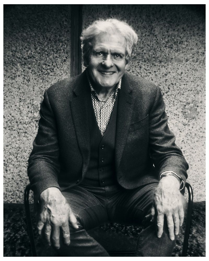 Monochrome portrait of an elderly man with white hair smiling while seated in a chair wearing a tailored blazer and patterned shirt.