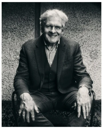 Monochrome portrait of an elderly man with white hair smiling while seated in a chair wearing a tailored blazer and patterned shirt.