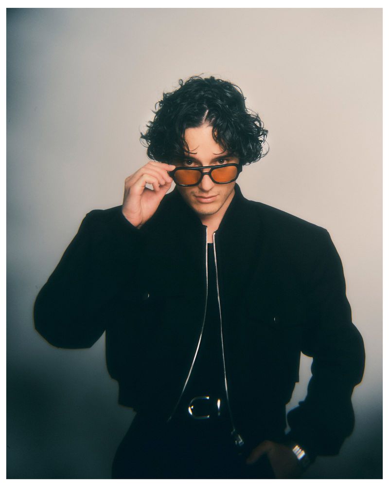 Actor Harry Collett wearing amber-tinted sunglasses and a black zip jacket against a neutral studio backdrop.