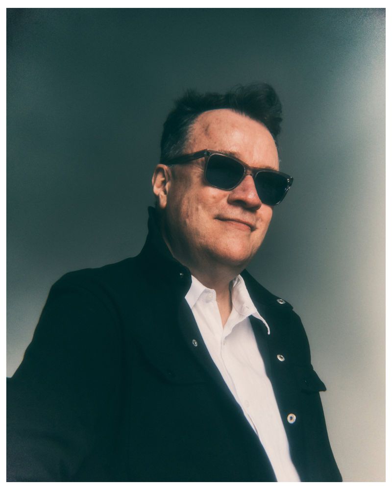 Man wearing dark sunglasses, a white collared shirt, and an open black jacket against a muted grey background.