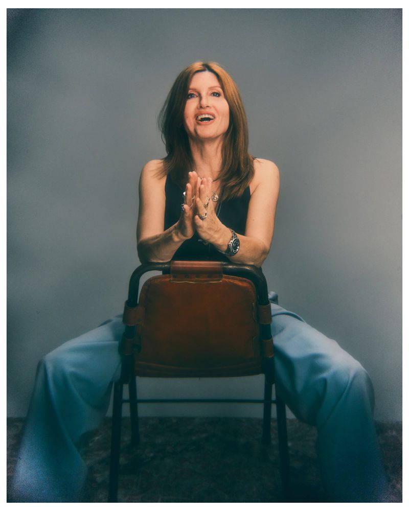 A smiling woman with auburn hair sits backwards on a brown leather chair wearing a black top and wide-leg light blue trousers.