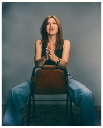 A smiling woman with auburn hair sits backwards on a brown leather chair wearing a black top and wide-leg light blue trousers.