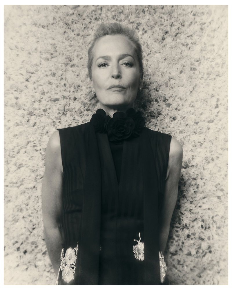 Monochrome portrait of actor Gillian Anderson wearing a sleeveless black garment with floral neck detail against a textured aggregate wall.