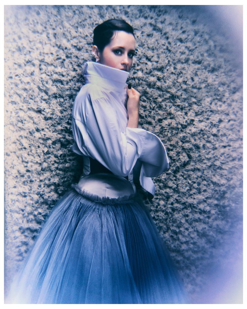 A woman with short dark hair in a high-collared white shirt and full blue tulle skirt poses against a textured backdrop.