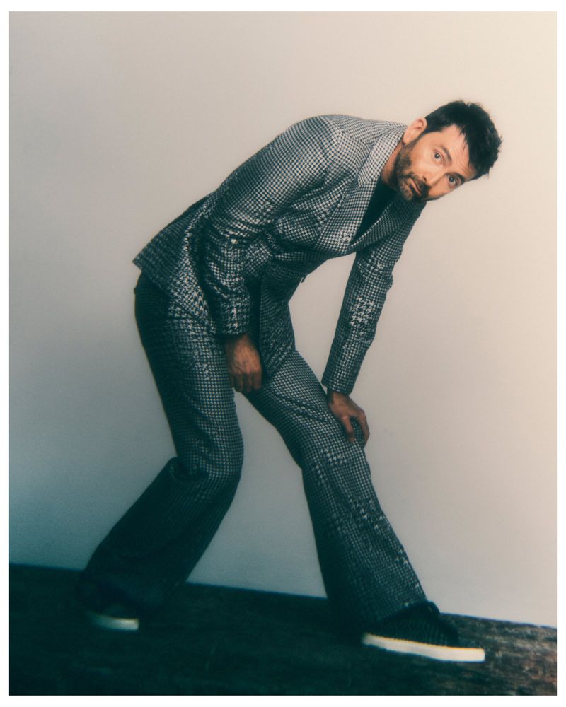 David Tennant in a grey patterned suit leaning forward against a studio wall with hands resting near knees.