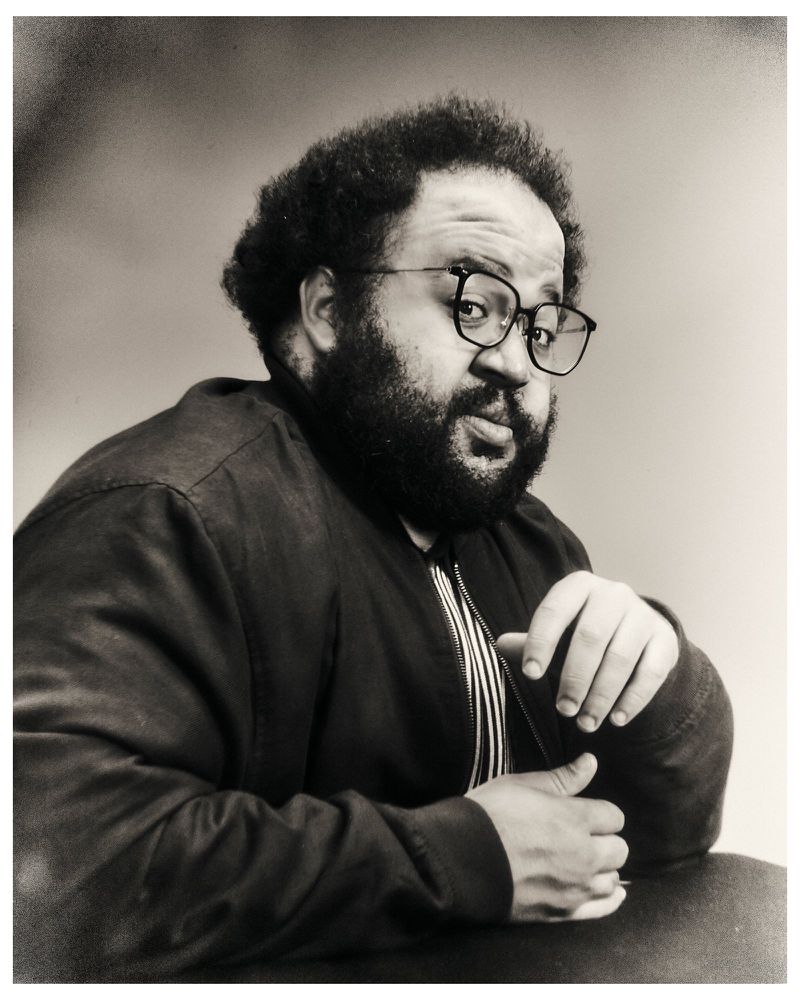 Monochrome portrait of a bearded man wearing black-rimmed eyeglasses and a dark jacket over a striped shirt against a grey backdrop.