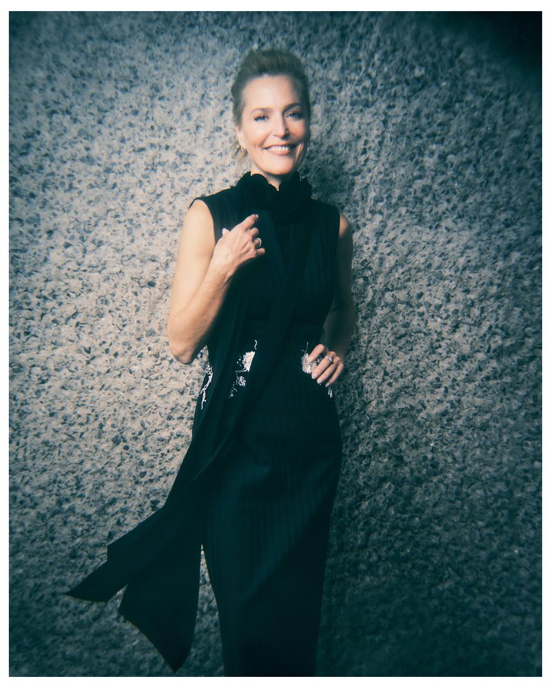 Actor Gillian Anderson smiling in a sleeveless black dress against a rough textured stone wall in an editorial portrait.