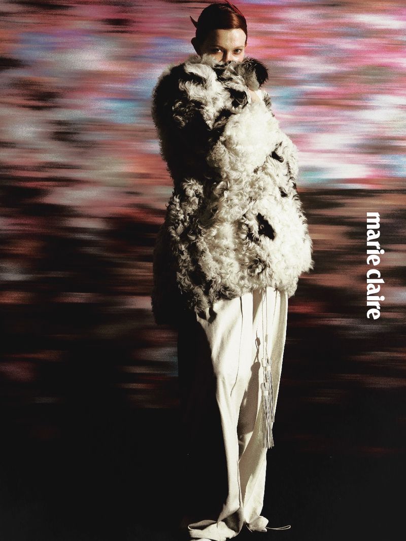 Model wearing a textured black and white faux-fur coat and light trousers against a blurred abstract background