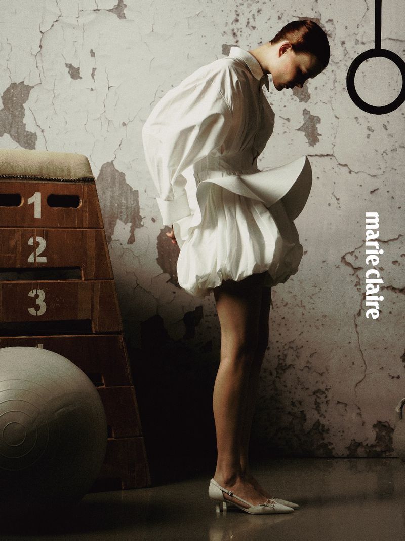 Model wearing a white structured dress and heels standing next to a wooden gym vault and exercise ball