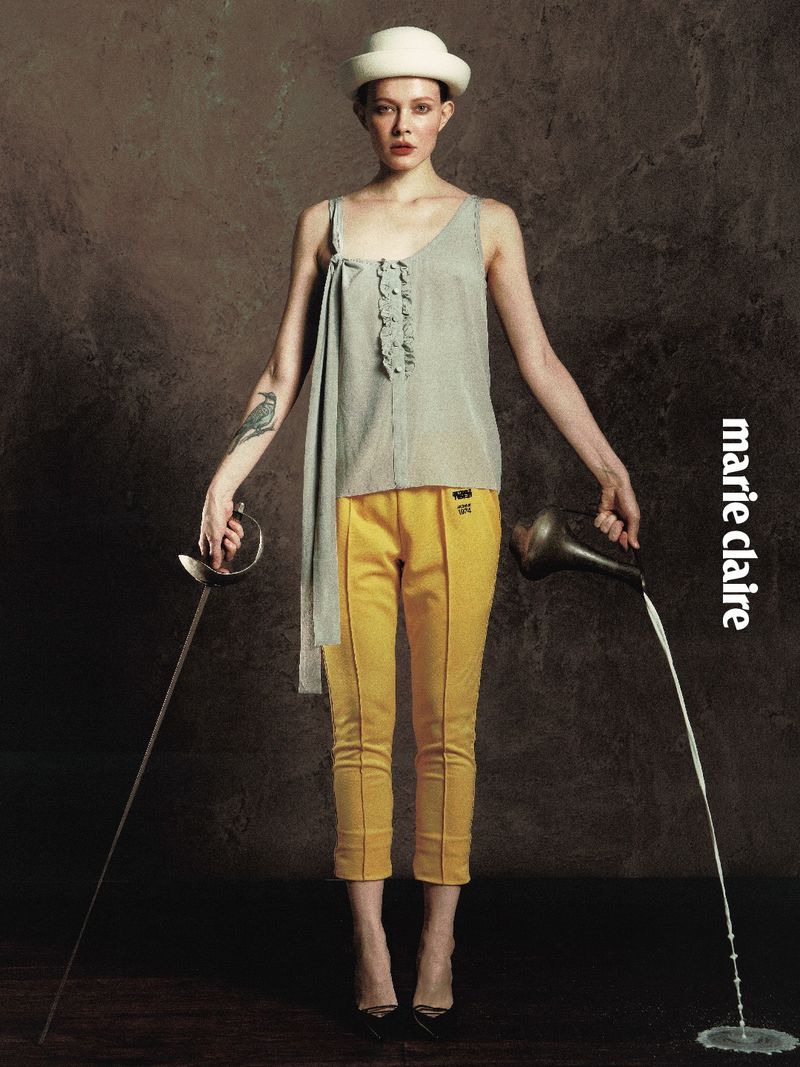 Model posing in yellow cropped trousers, a grey draped top, and a white hat while holding a fencing foil and pouring water from a pitcher.