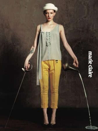 Model posing in yellow cropped trousers, a grey draped top, and a white hat while holding a fencing foil and pouring water from a pitcher.