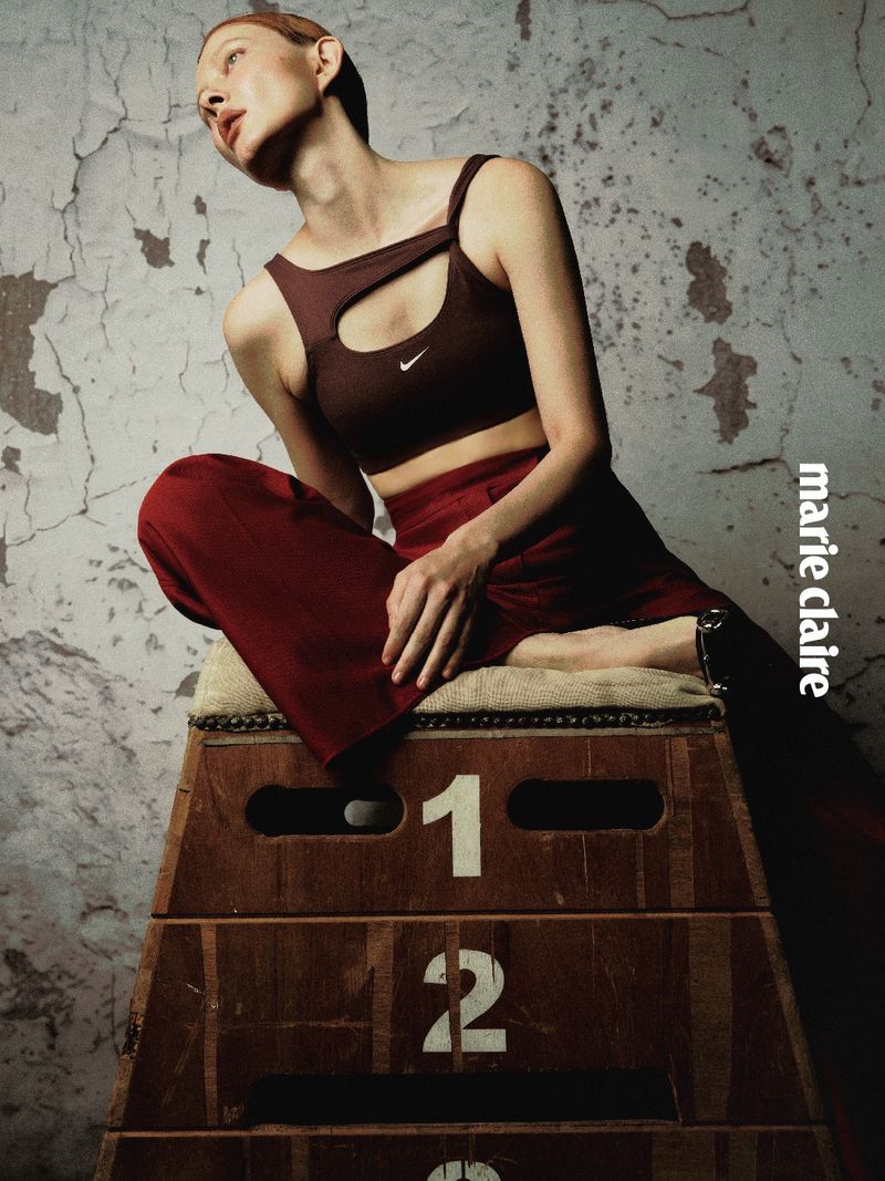 Model Sabi Khazgaleeva wearing a burgundy cutout sports top and pants, seated atop a numbered wooden gymnastics vaulting box.