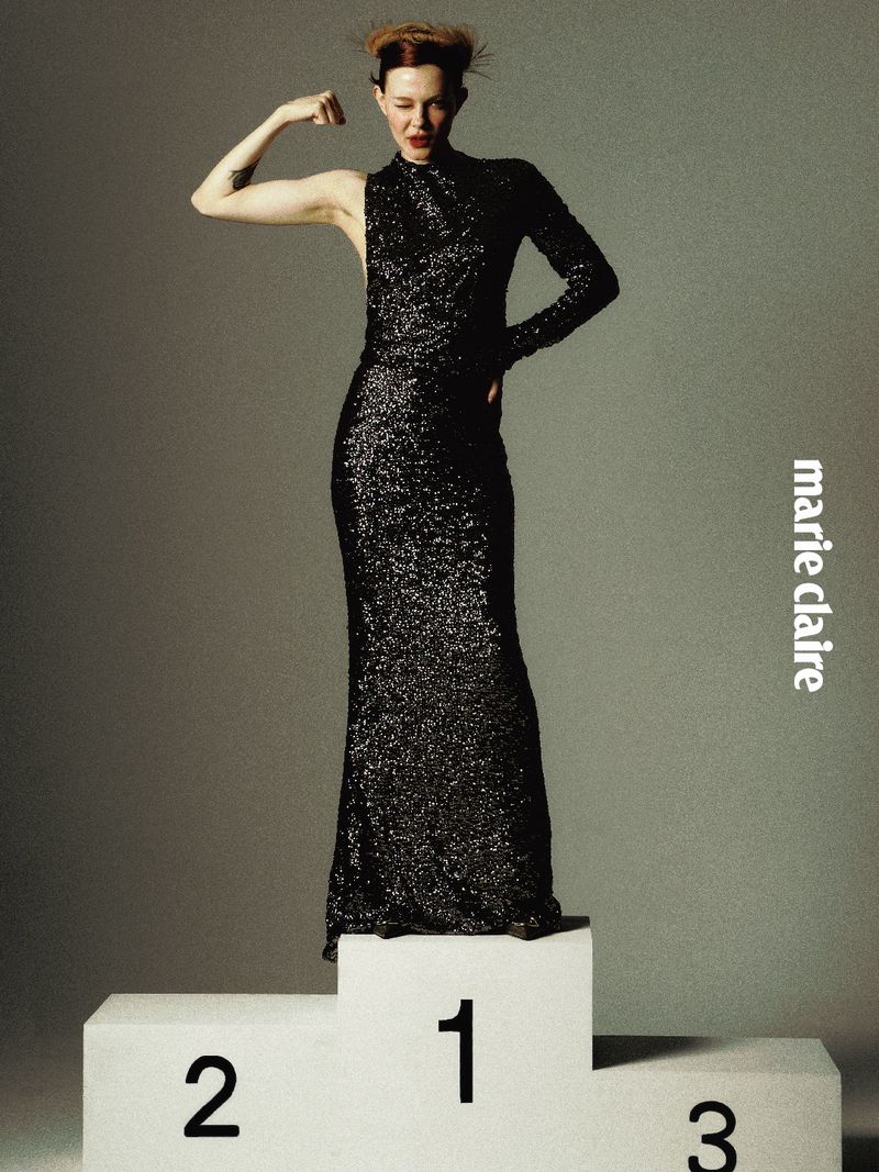 Model posing in a black sequin asymmetric gown on a numbered winners podium against a grey background.