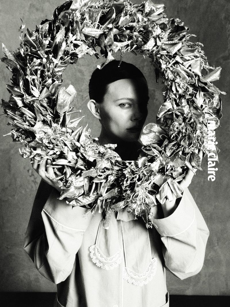 Model Sabi Khazgaleeva holds a large textured metallic wreath framing her face in a black and white editorial portrait.