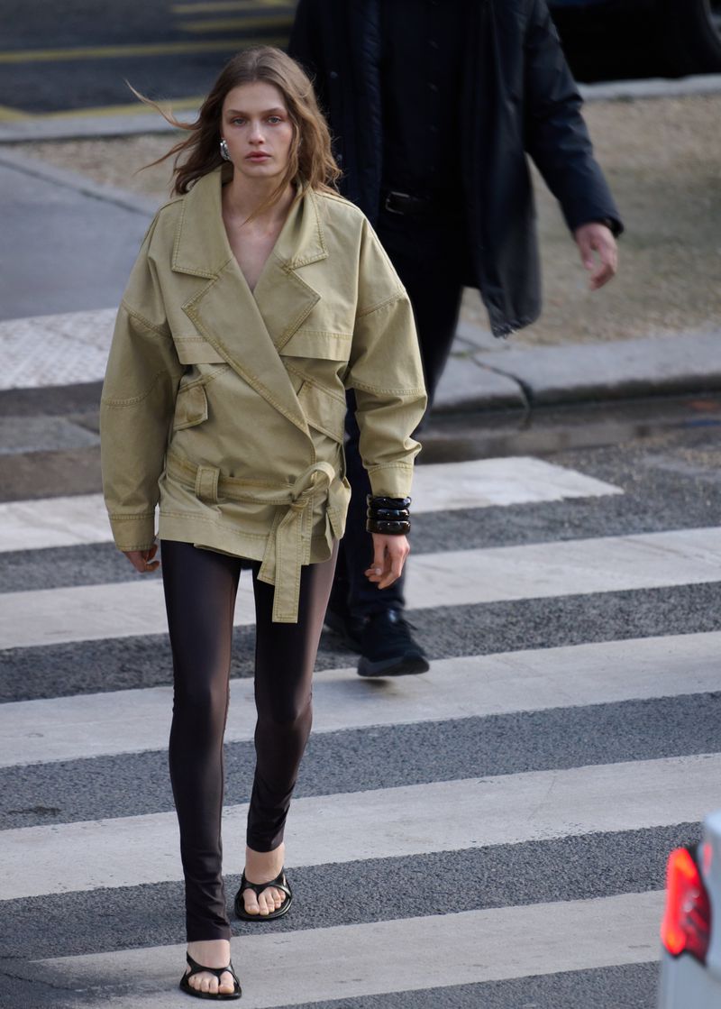 Model Ella Mccutcheon crossing a city street wearing a belted khaki trench jacket, dark leggings, and black heeled sandals.