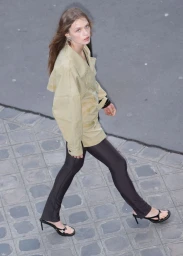 Model Ella Mccutcheon walks across a paved street in a beige oversized jacket, black leggings, and black kitten-heel thong sandals.