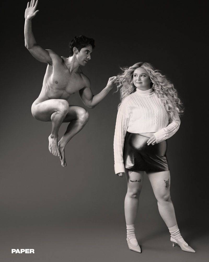 Megan Stalter standing in a white knit sweater beside a naked man suspended mid-air in a monochrome studio portrait.