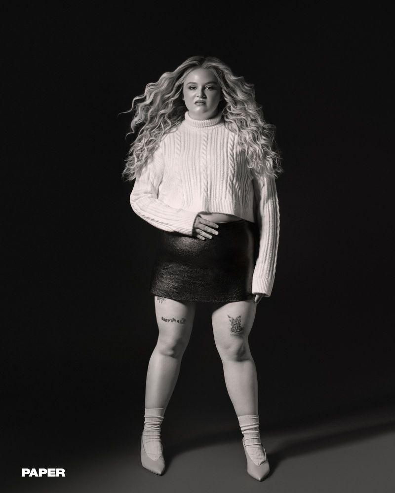Megan Stalter poses in a cropped white cable-knit sweater and black mini skirt against a dark studio background for Paper Magazine.