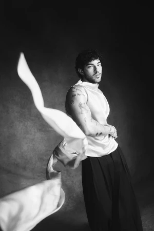 Black and white portrait of Luis Capecchi wearing a draped sleeveless top and trousers in front of a textured backdrop.