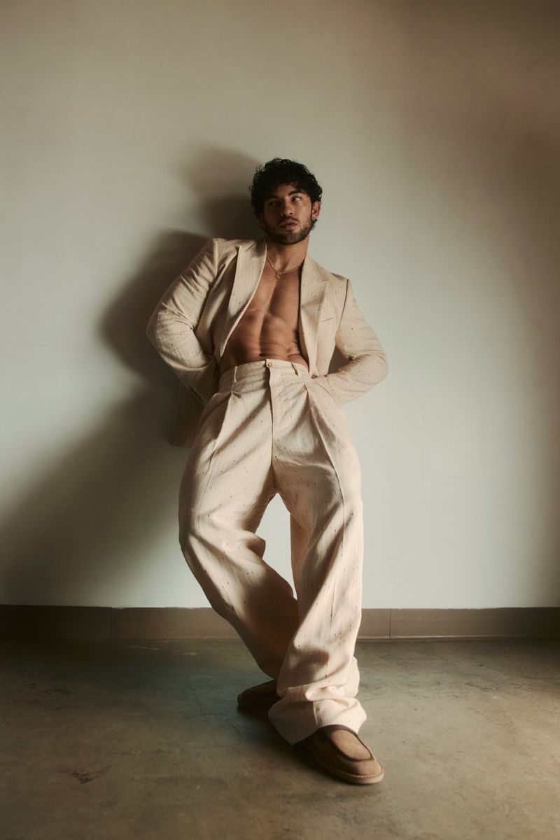 Luis Capecchi posing shirtless in an open cream tailored suit with wide-leg trousers leaning against a neutral wall in warm light.