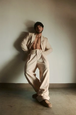 Luis Capecchi posing shirtless in an open cream tailored suit with wide-leg trousers leaning against a neutral wall in warm light.