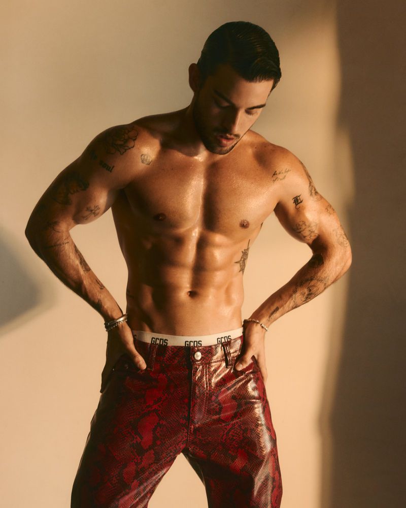 Shirtless tattooed man posing in glossy red snakeskin trousers with white branded waistband against a warm neutral studio backdrop.