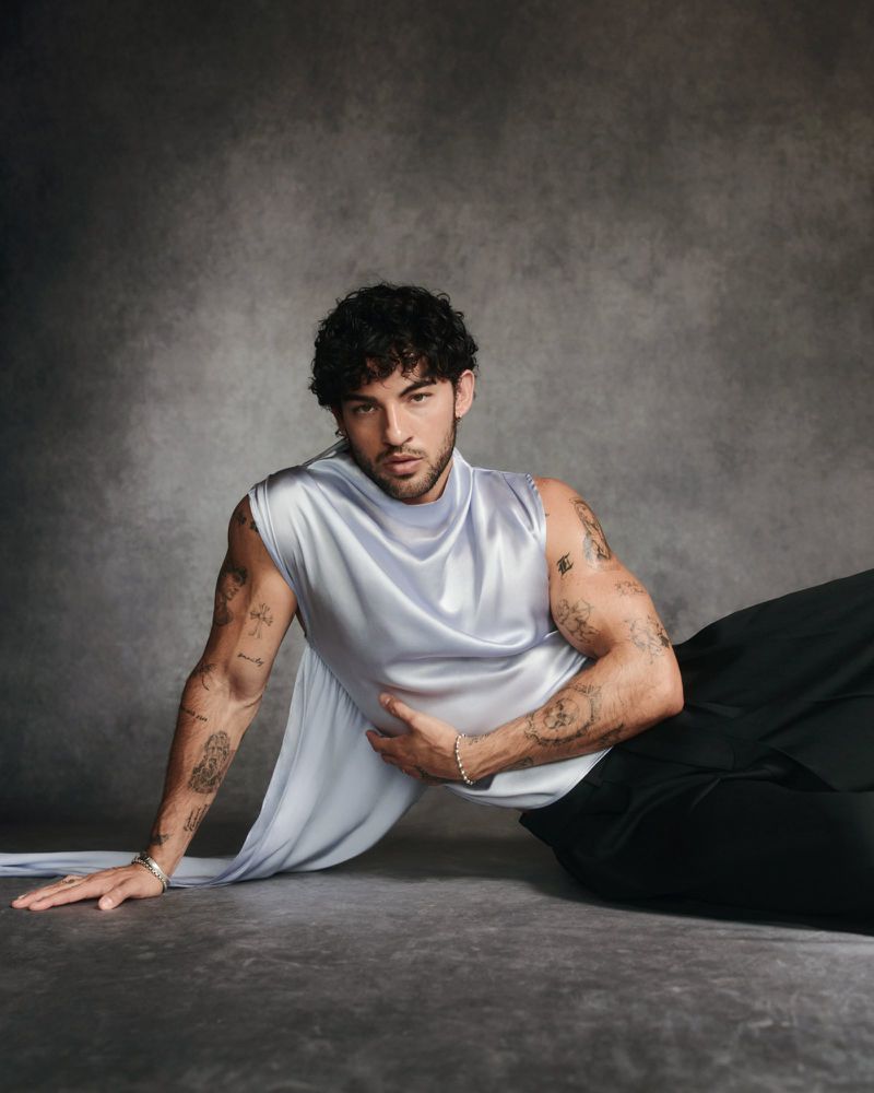 Luis Capecchi reclines in a sleeveless draped lilac satin top and black trousers against a mottled grey studio backdrop.
