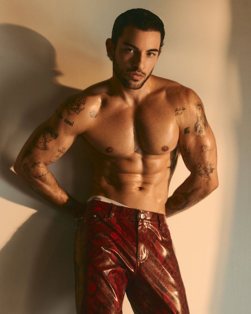 Shirtless man with tattoos wearing red patterned trousers against a warm lit wall