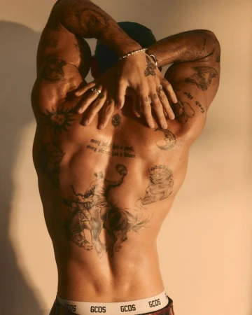 Rear view of shirtless model Luis Capecchi with hands behind his head, showing his back tattoos against a warm studio backdrop.