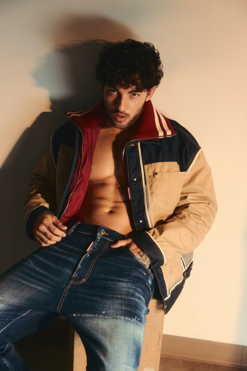 Luis Capecchi poses shirtless in an open colorblocked jacket and blue jeans against a neutral wall.