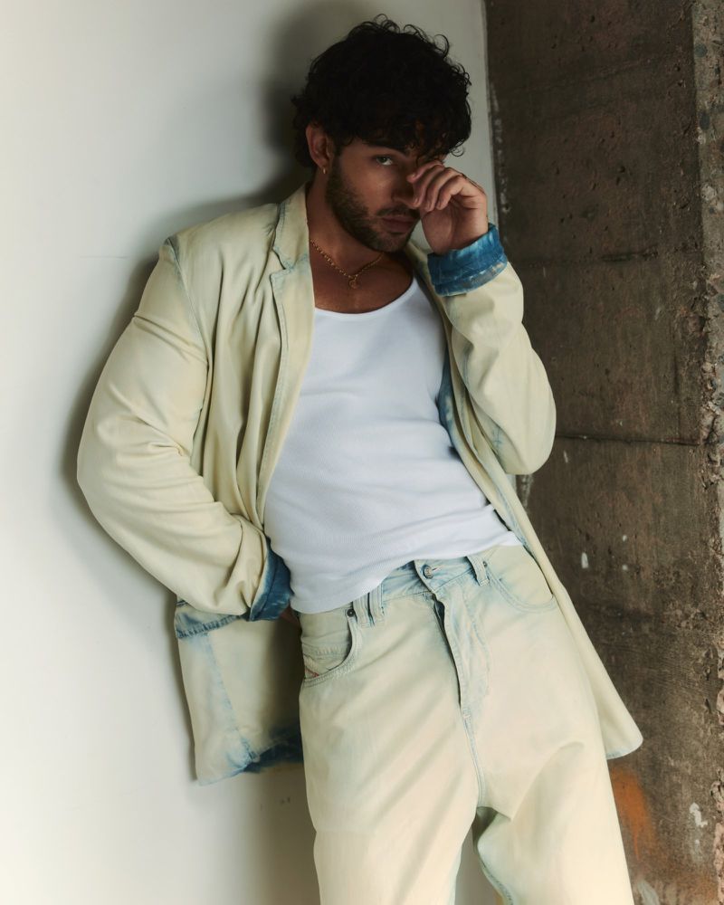 Luis Capecchi wearing a pale washed denim jacket, white tank top, and light jeans leaning against a concrete wall.