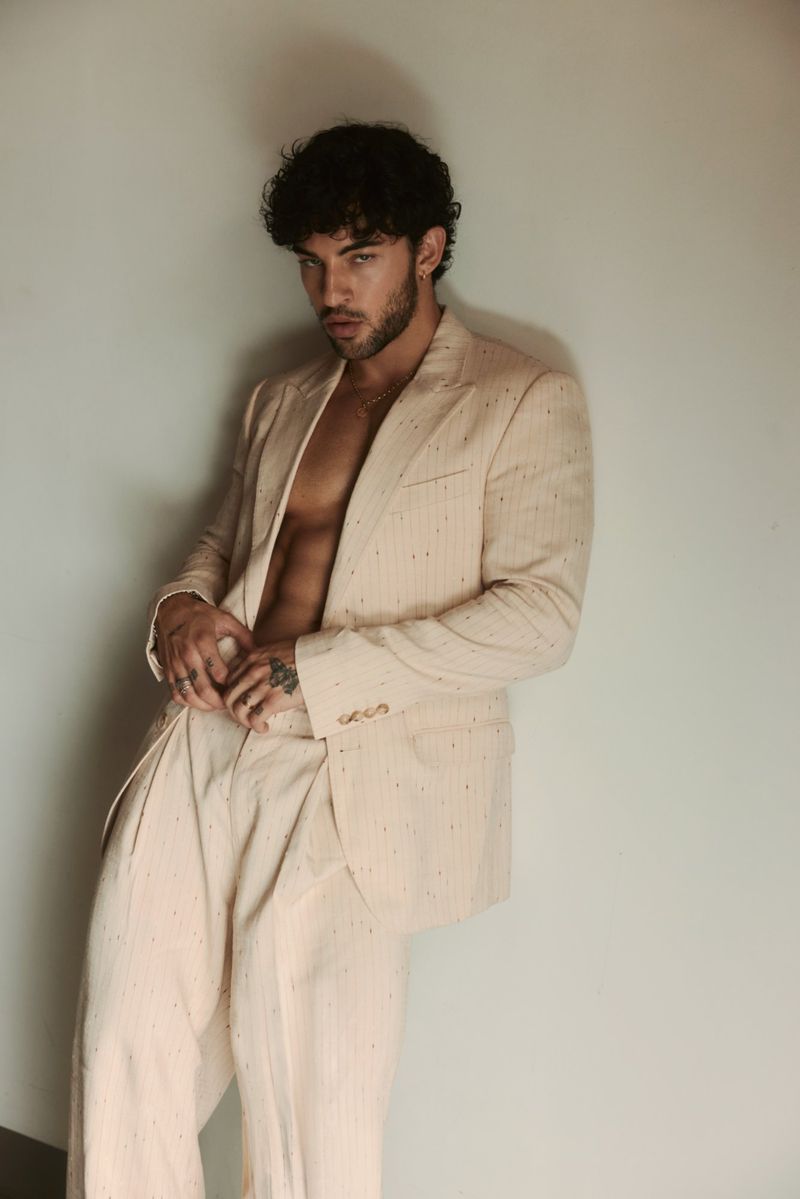 Luis Capecchi poses shirtless in a cream tailored suit against a neutral studio backdrop.