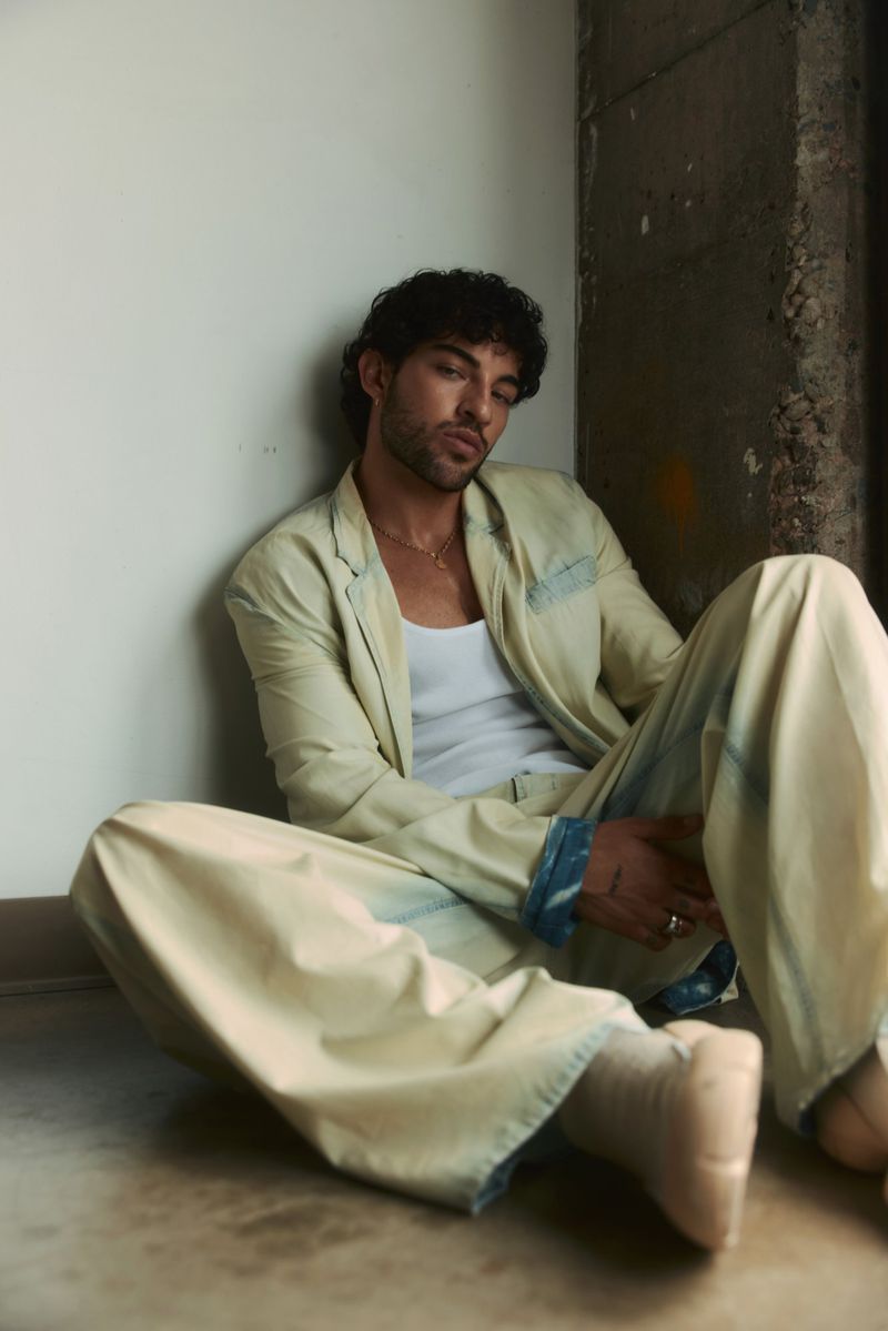Luis Capecchi wearing an oversized pale yellow suit and white tank top, seated on the floor leaning against an interior wall.