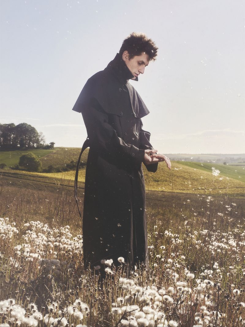 Model wearing a long black coat standing in a rural meadow filled with white dandelion seed heads under soft sunlight.