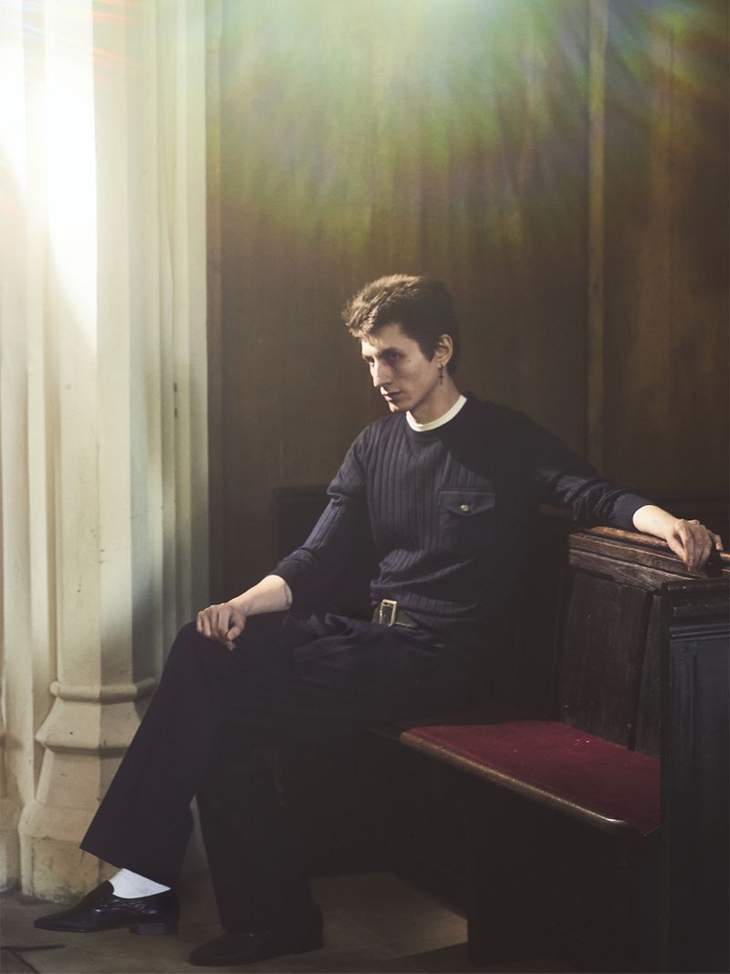 Model Henry Kitcher seated on a wooden church pew wearing a dark ribbed sweater, dark trousers, and white socks.