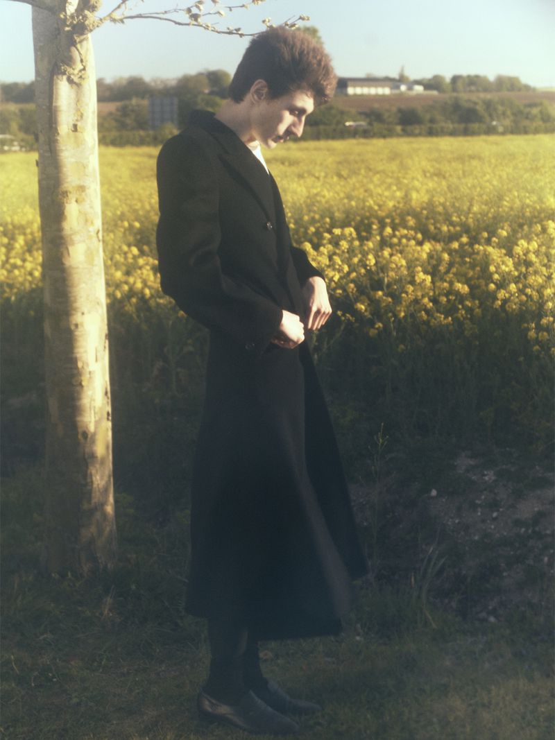 Model Henry Kitcher stands in profile wearing a long black tailored coat beside a tree trunk near a yellow flowering field.