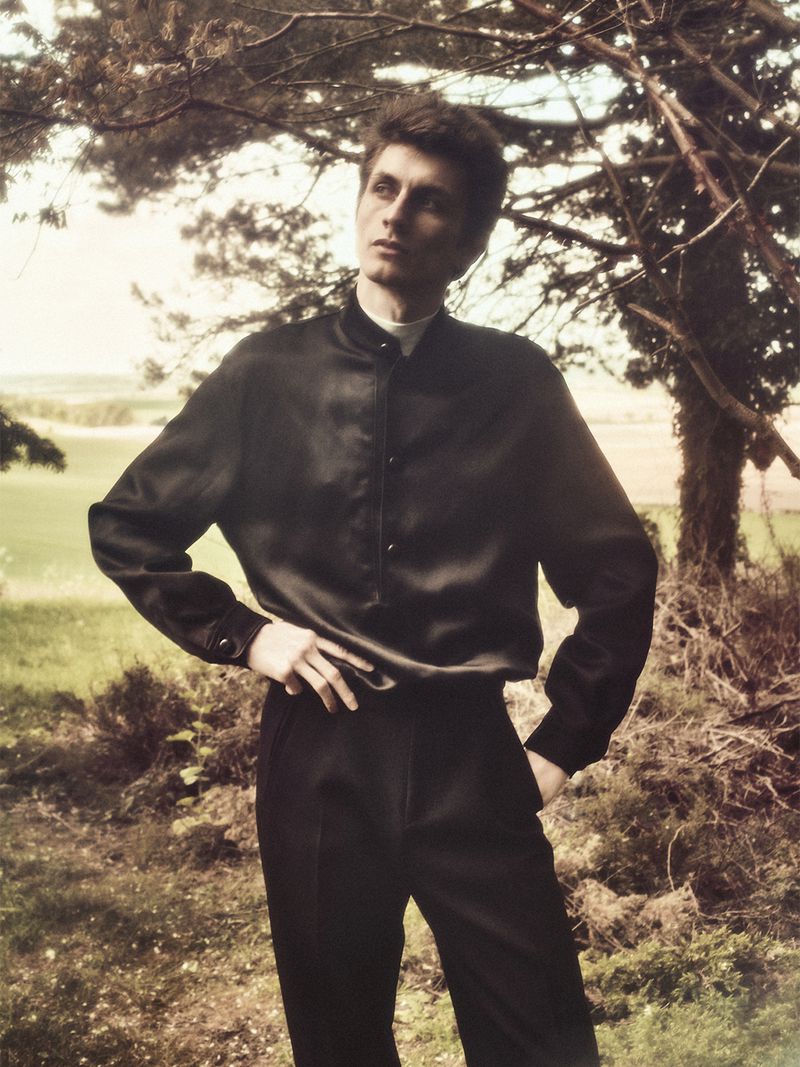 Henry Kitcher poses outdoors under tree branches wearing a black satin button-up shirt and dark trousers for The Vale.
