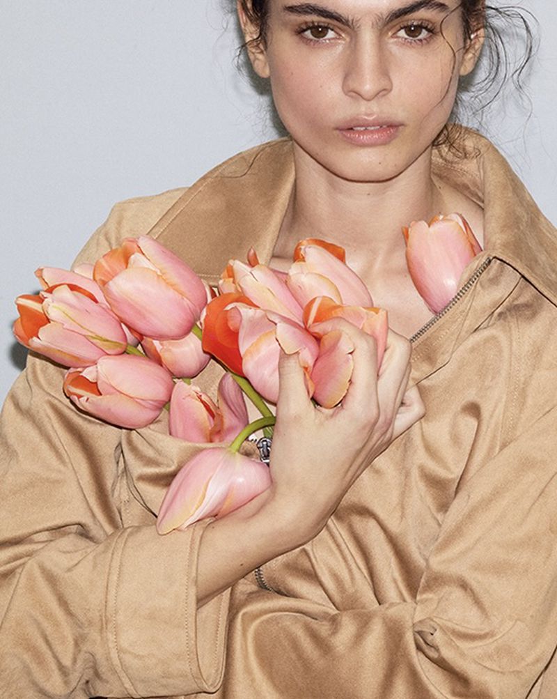 Close-up of a model wearing a tan suede jacket holding a cluster of fresh pink and peach tulips against a neutral studio backdrop.