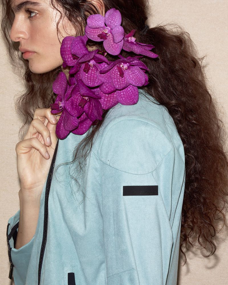 Model Carol Monteiro wears purple orchids styled around her neck with a pale blue zip jacket against a neutral studio backdrop.