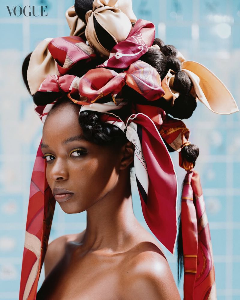 Portrait of a Black woman with an elaborate sculptural headwrap made of red and cream patterned silk scarves against blue tiles.