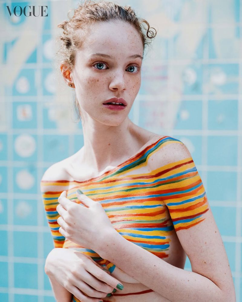 Woman with freckles and colorful striped body paint across her chest and shoulders posing before a light blue tiled backdrop.