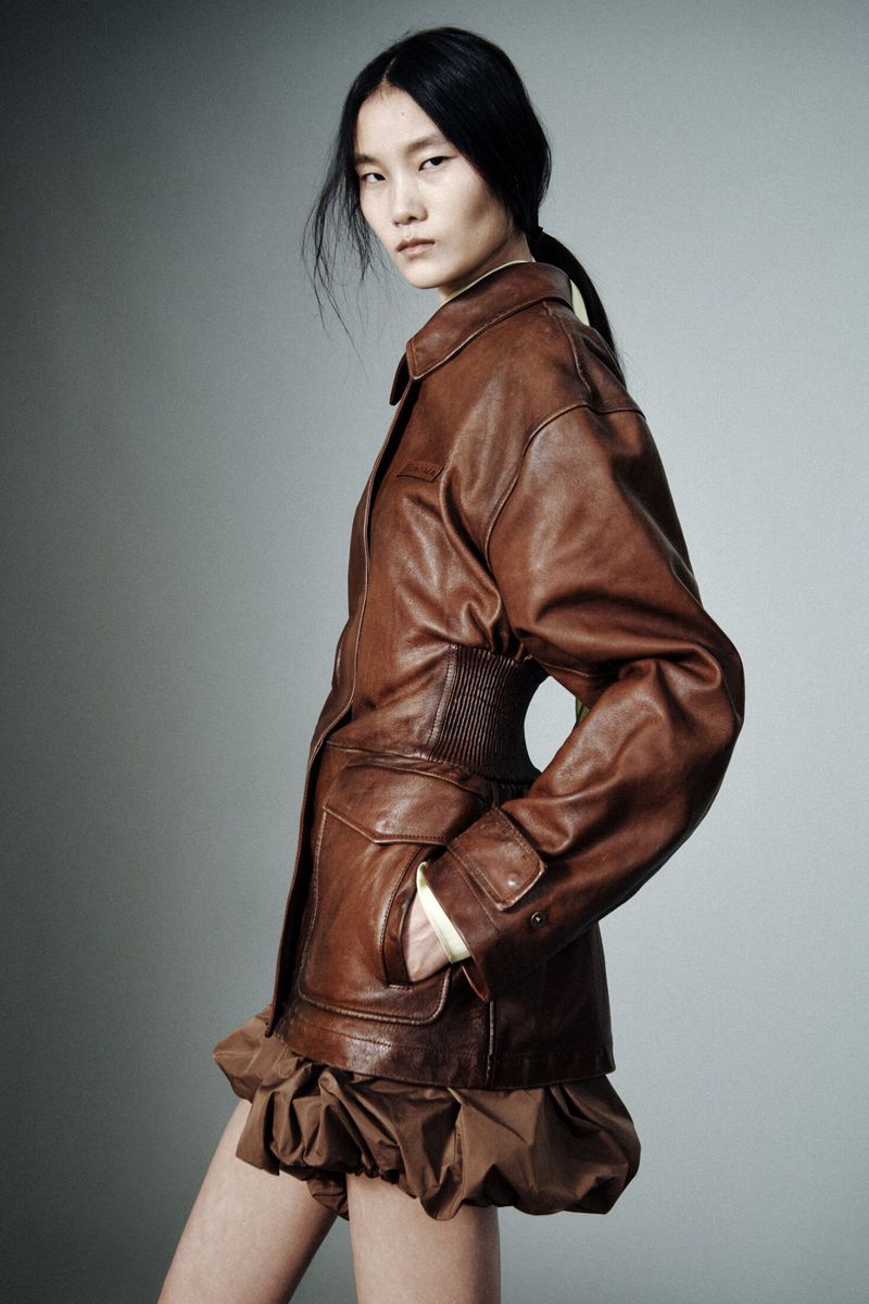 Model in a cinched brown leather jacket and gathered bubble hem skirt posing in profile against a grey studio backdrop.