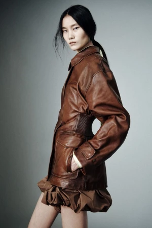 Model in a cinched brown leather jacket and gathered bubble hem skirt posing in profile against a grey studio backdrop.